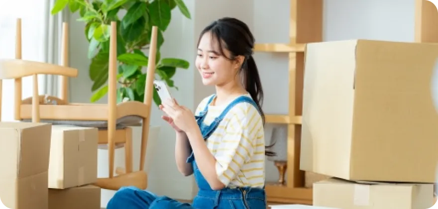 イメージ写真：引っ越しの段ボール箱に囲まれてスマホを操作している女性が写っています